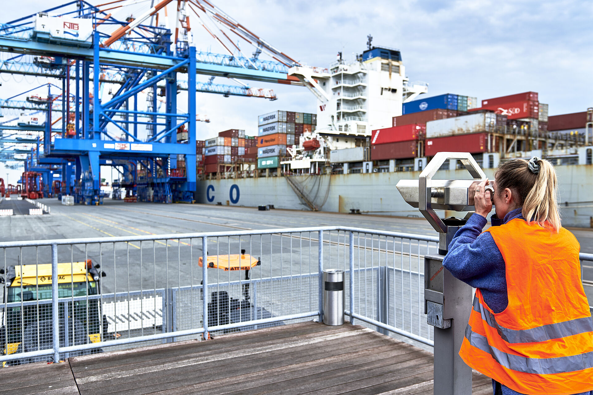 Frau mit Warnweste, rechts im Bild, schaut vom Aussichtspunkt durch ein Fernglas auf das Containerterminal, rechts im bild