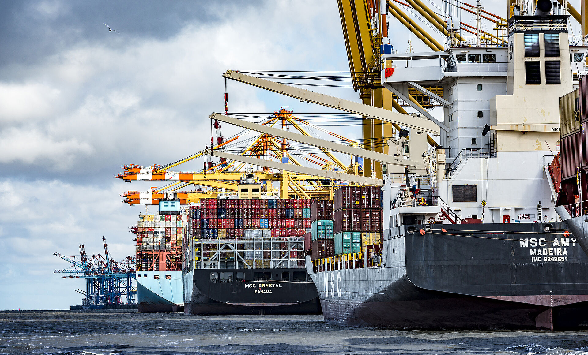 Das Containerterminal Bremerhaven in der rechten Bildhälfte. Kranarme und Containerbrücken überragen 4 Schiffe, die dort liegen. Leichte Bewölkung.