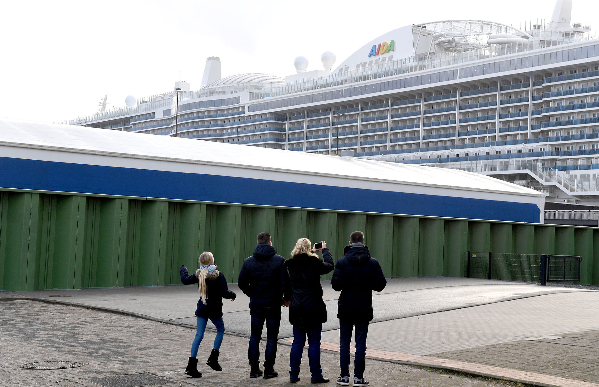Im Vordergrund eine Gruppe Menschen/Familie, die ein Aida-Kreuzfahrtschiff fotografieren, links im Mittelgrund eine grüne Spundwand.
