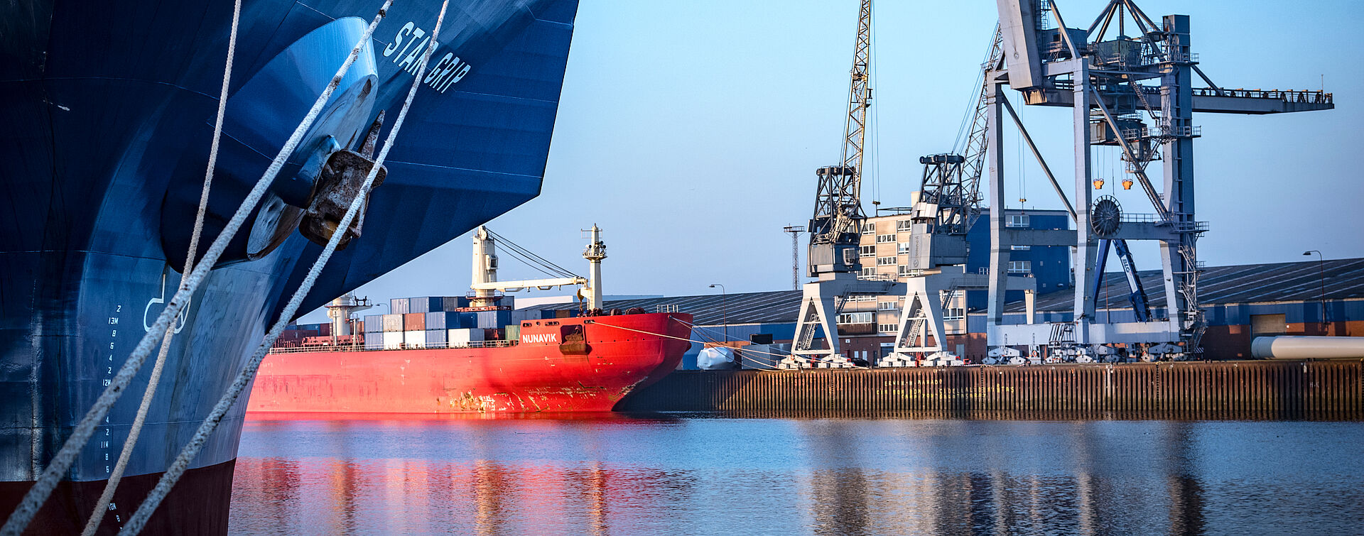 Ein Schiff mit rotem Unterschiff liegt im Hafenbecken an der Kaje. rechts Kräne oder Brücken.