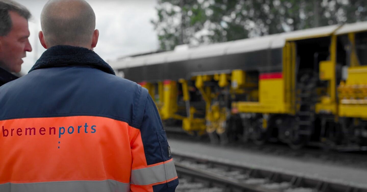 Bremenportsmitarbeiter mit Jacke in Neonorange am Gleis vor gelbem Zug.