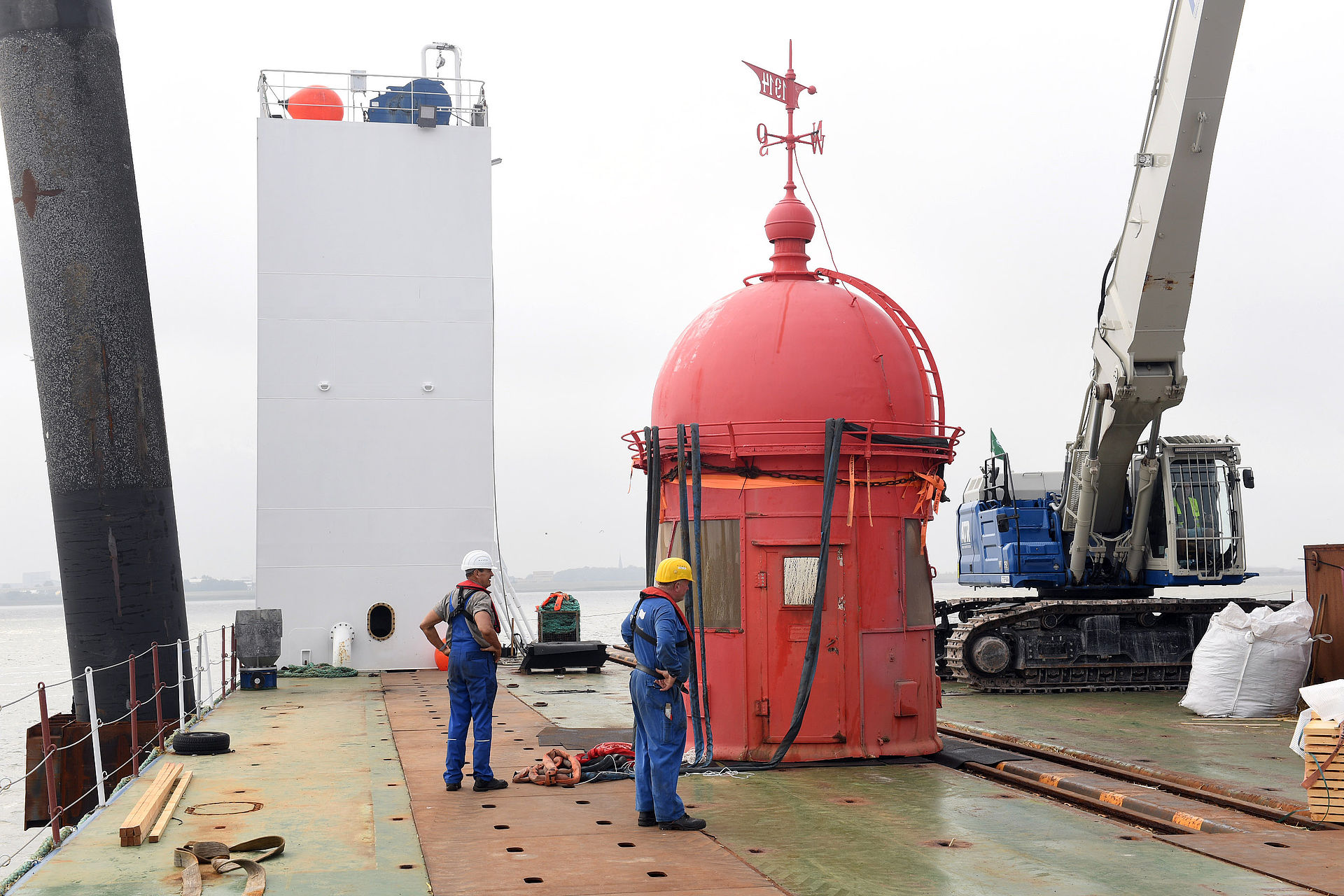 Die rote Kuppel des Molenturms auf Deck eines Schiffes. Davor Arbeiter mit gelben Helmen