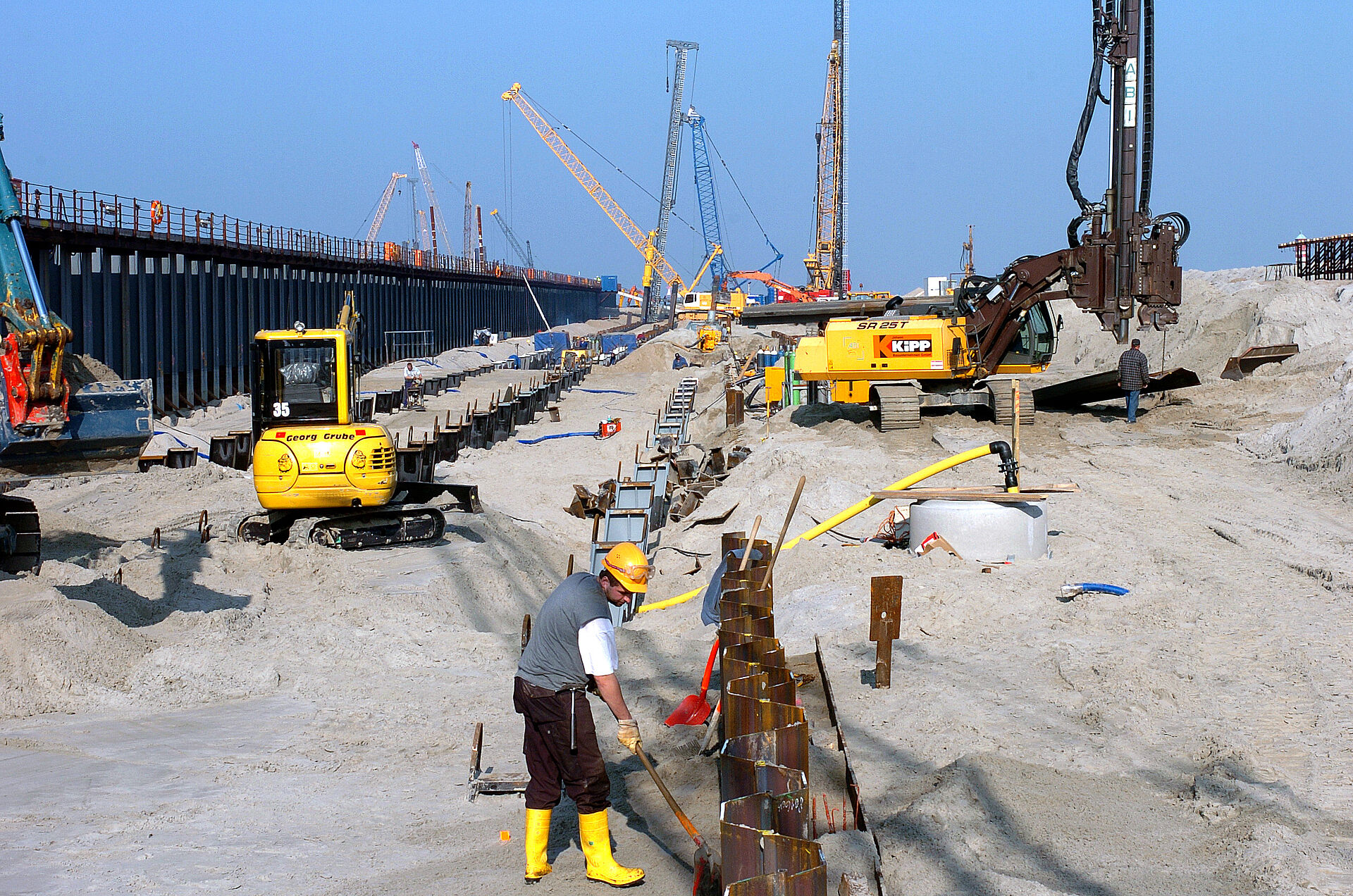 Ein Blick auf die Baustelle der Erweiterung Columbuskaje. Im Vordergrund ein Bauarbeiter mit gelbem Helm und Stiefeln. Im Hintergrund Kräne. Davor schauen die Köpfe einer Reihe Stahlprofile aus dem Sand.