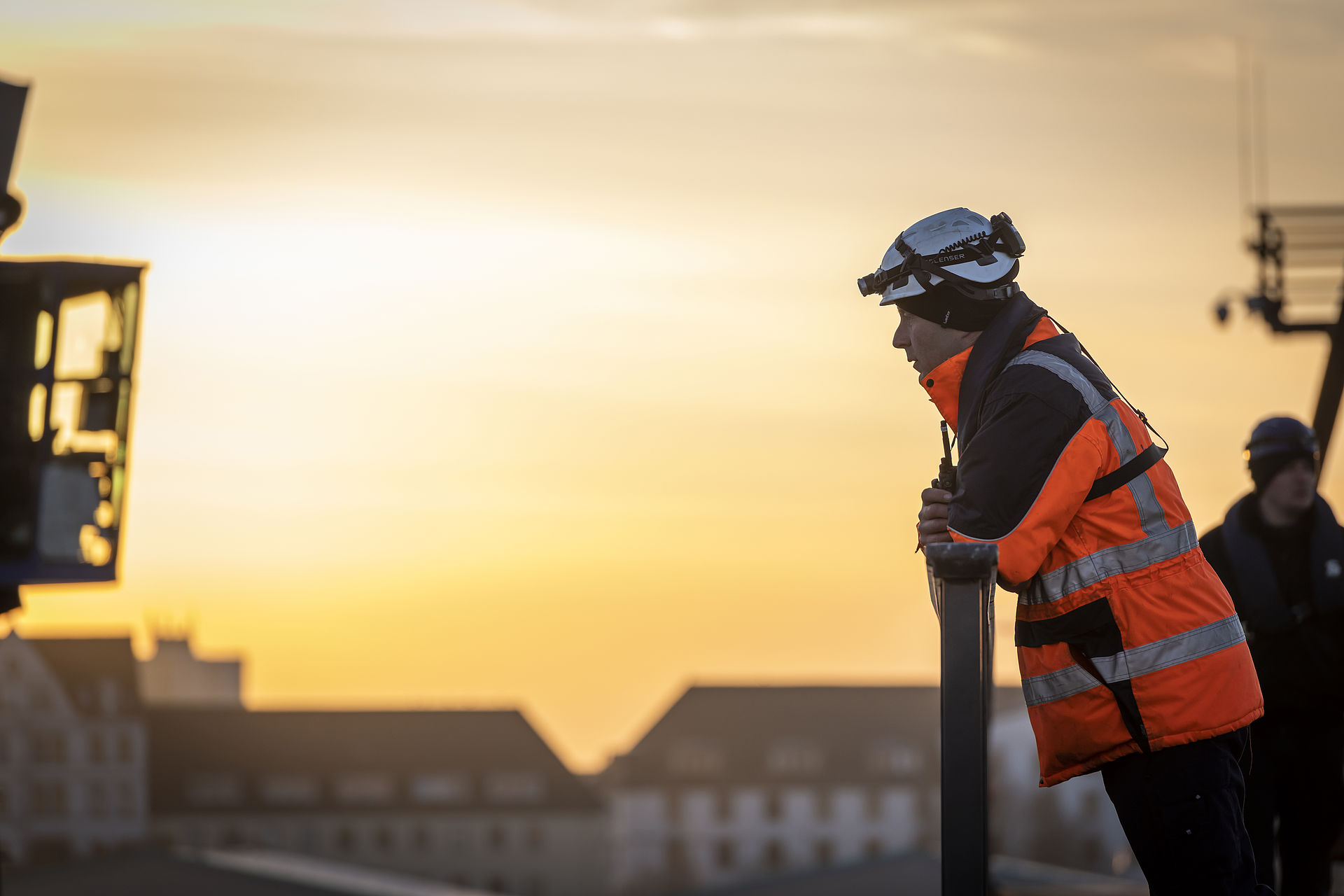 Mitarbeiter von Bremenports mit Helm, Stirnlampe und Funkgerät blickt nach Links, im Hintergrund Sonnenaufgang