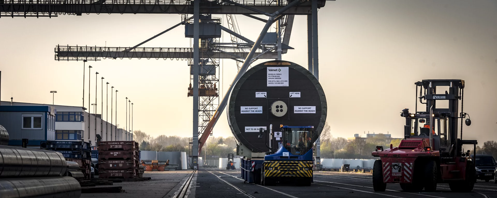 In der Mitte des Bildes steht ein großes zylindrische Objekt, das transportiert wird. Kräne, Lagerhallen und ein Gabelstapler sind ebenfalls zu sehen.