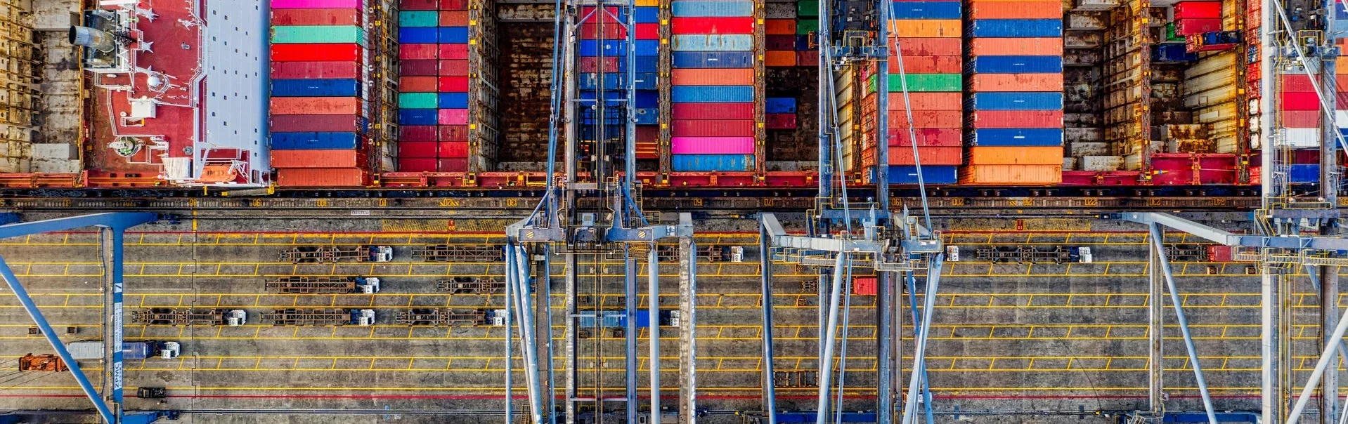 Luftaufnahme vom Betriebsauflauf eines Hafens. Containern sind aufeinander gestapelt, Kräne und verschiedene Fahrzeuge befinden sich auf dem Gelände.